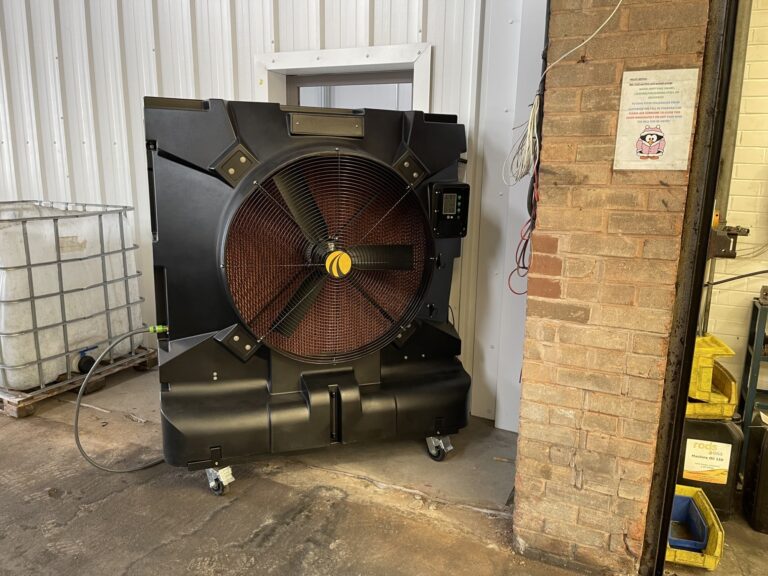 AlphaPrecision Large industrial fan on wheels next to a brick wall and a white container in a warehouse.